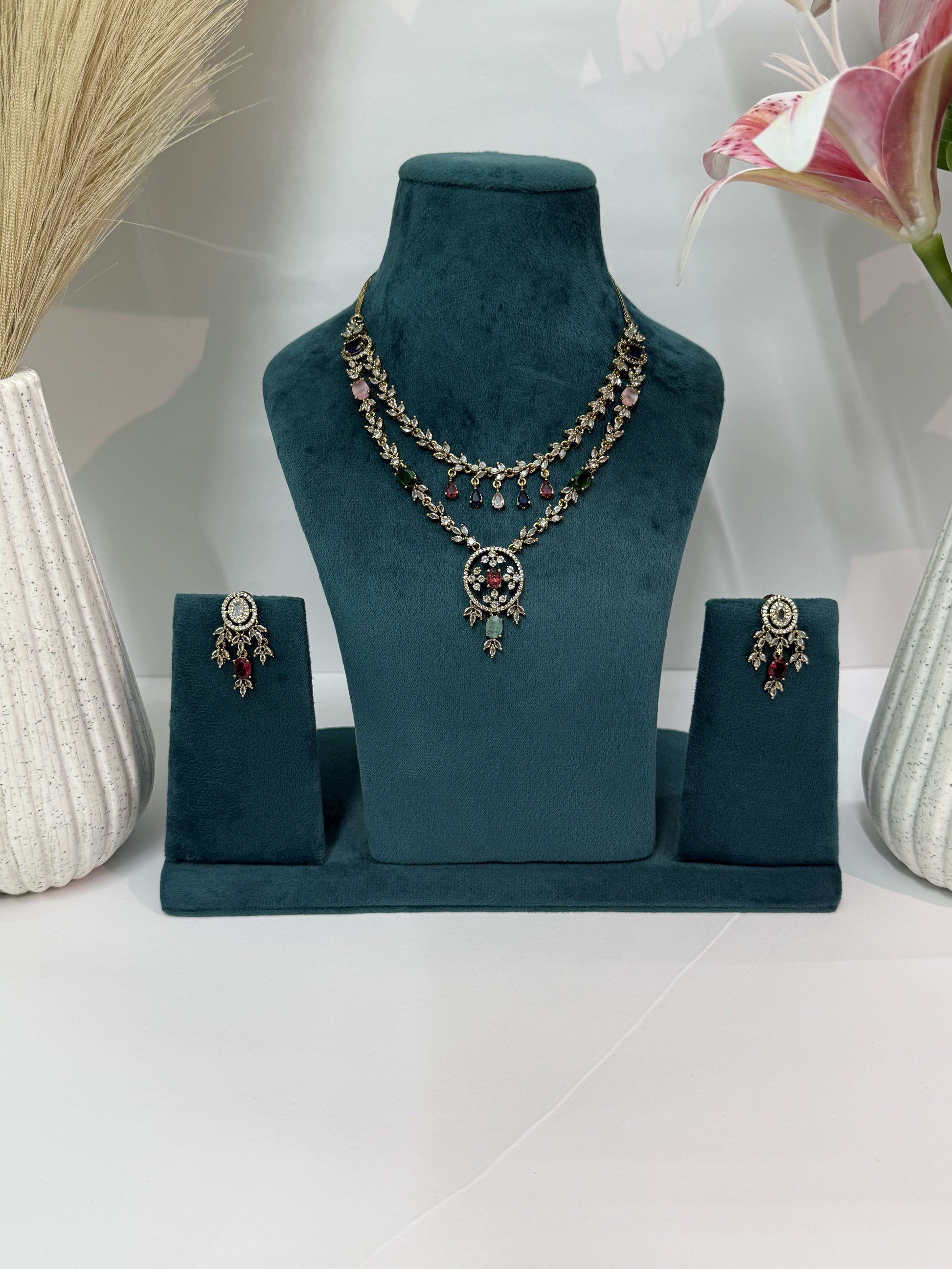 A gold-toned jewelry set displayed on a dark teal velvet stand, featuring a layered necklace with a circular pendant and matching drop earrings, styled with decorative dried grass and a pink flower in white ribbed vases.