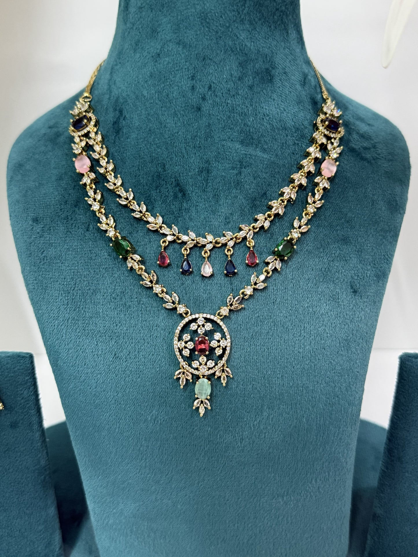 A double-strand gold-toned necklace with colorful gemstone accents, displayed on a teal velvet jewelry bust. The necklace features gold leaf-patterned chains, pink and green stones, and a central circular pendant with a red heart and light green teardrop.