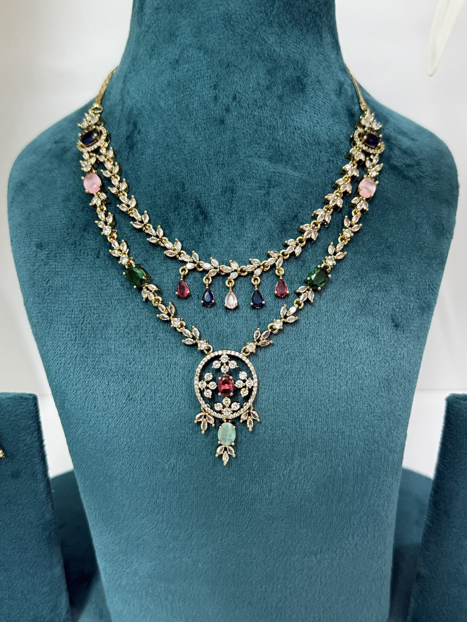 A double-strand gold-toned necklace with colorful gemstone accents, displayed on a teal velvet jewelry bust. The necklace features gold leaf-patterned chains, pink and green stones, and a central circular pendant with a red heart and light green teardrop.
